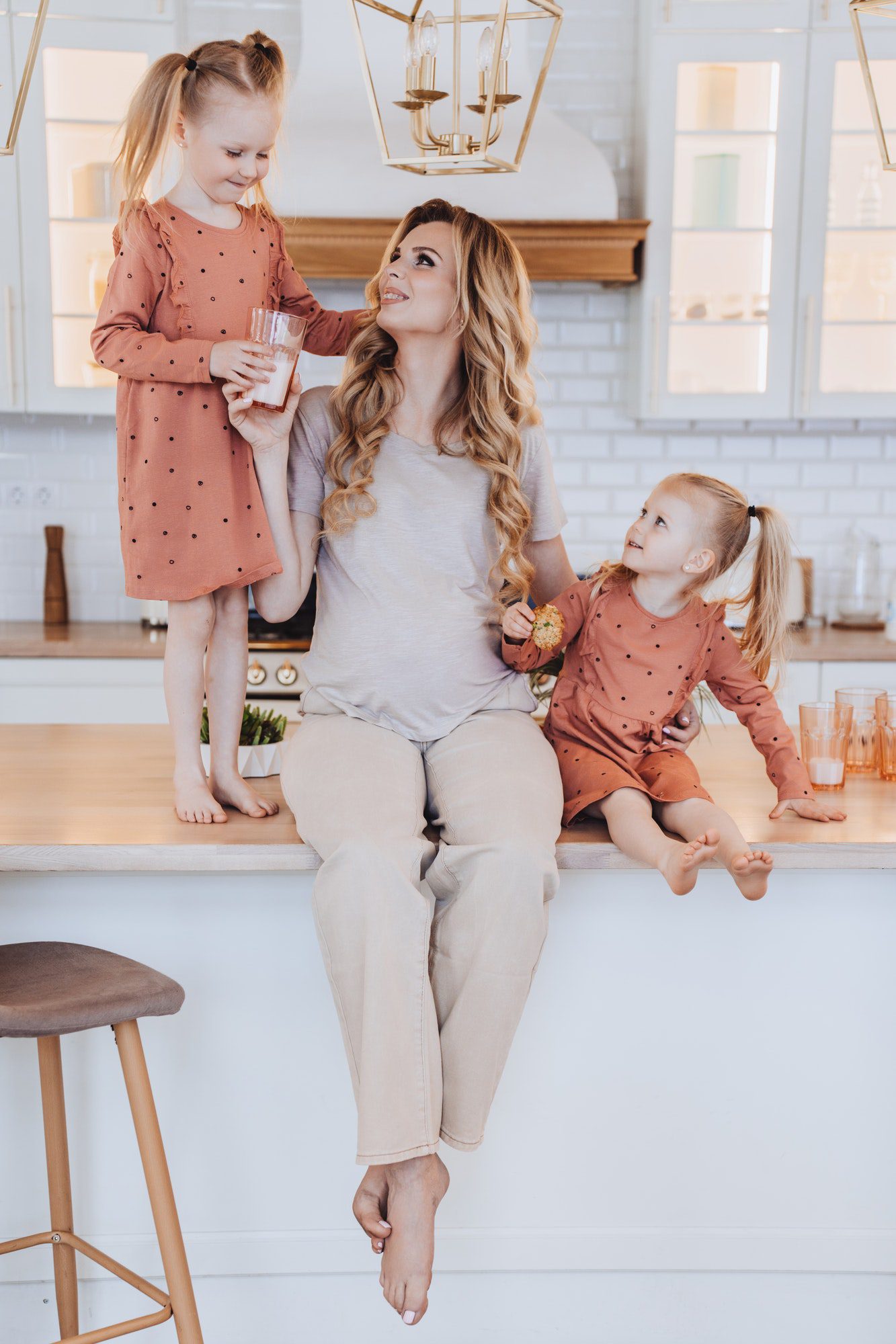 Pregnant caucasian mom sits on kitchen table with little daughters dressed in beige casual clothes.